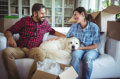 man and woman sitting with dog on couch surrounded by moving boxes